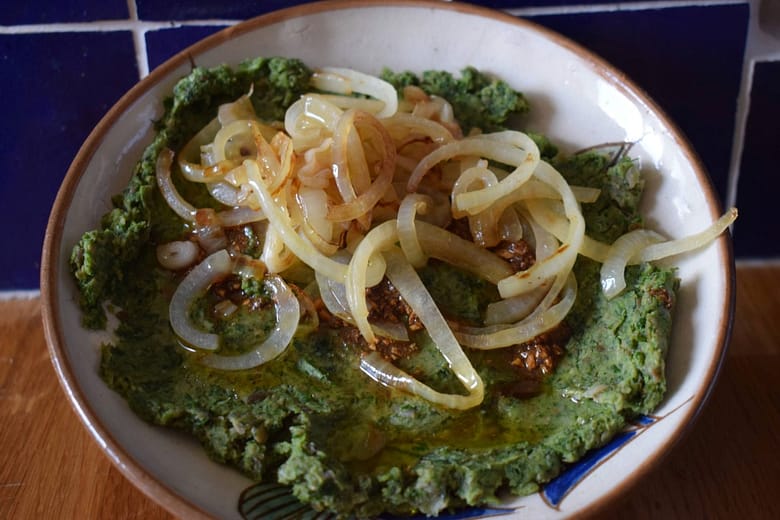 A bowl of Egyptian bissara made from mashed fava beans, topped with fried onions and spiced olive oil, served in a shallow ceramic dish.
