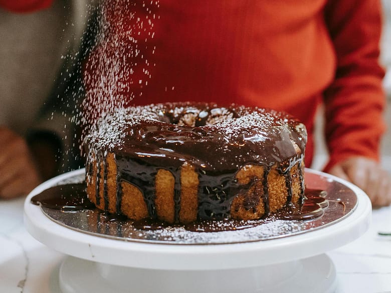 A golden cake topped with chocolate glaze being dusted with powdered sugar.