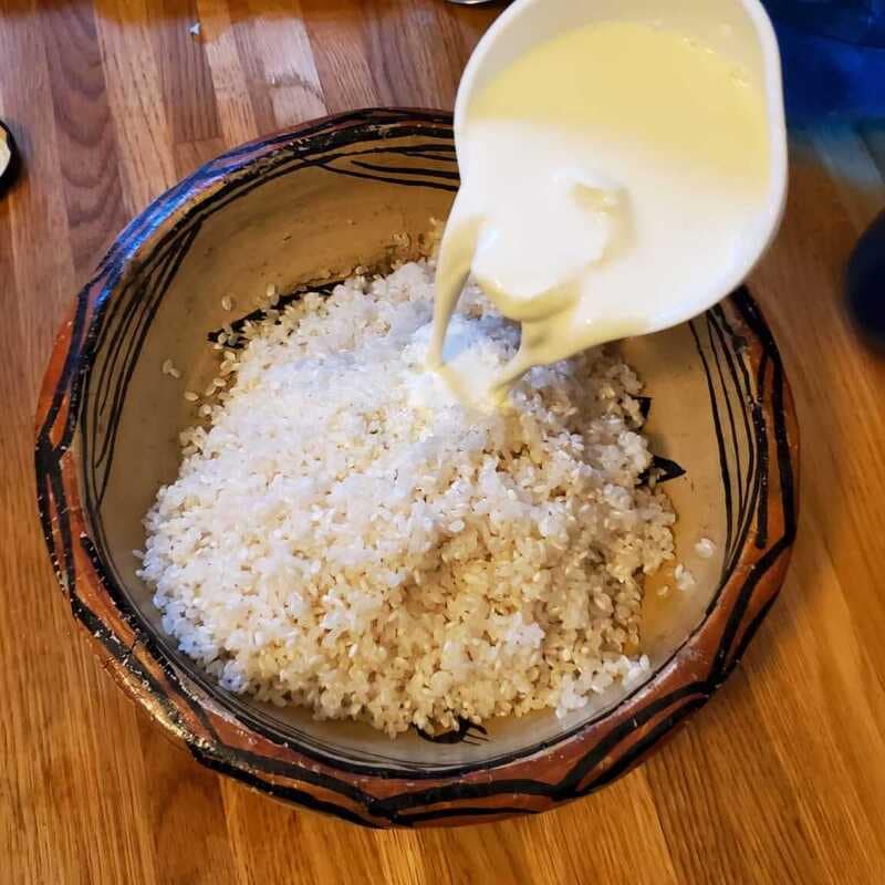 Milk being poured from a white pitcher over a mound of uncooked white rice in a traditional clay pot.